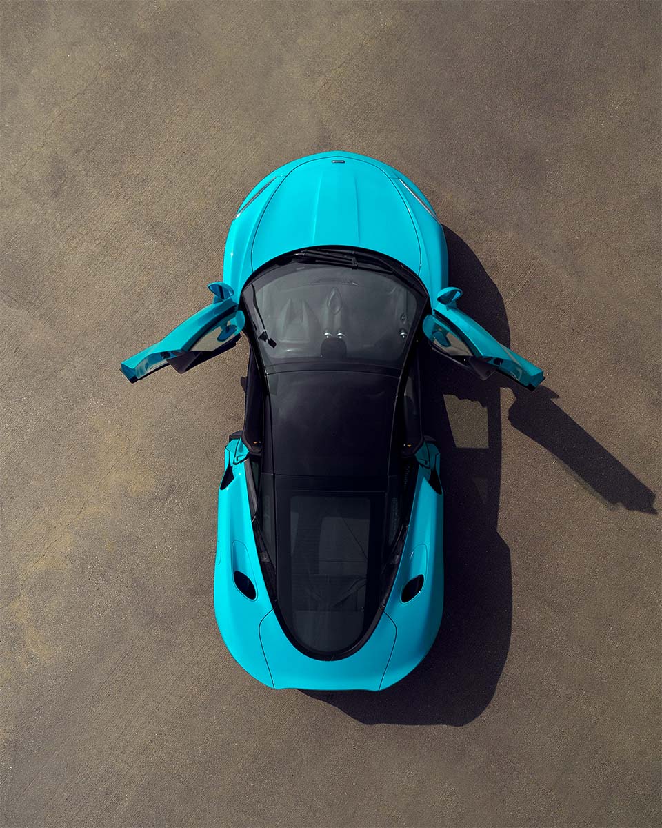 Top-down aerial of a cyan McLaren GT with its dihedral doors open on concrete, by Niaz Uddin
