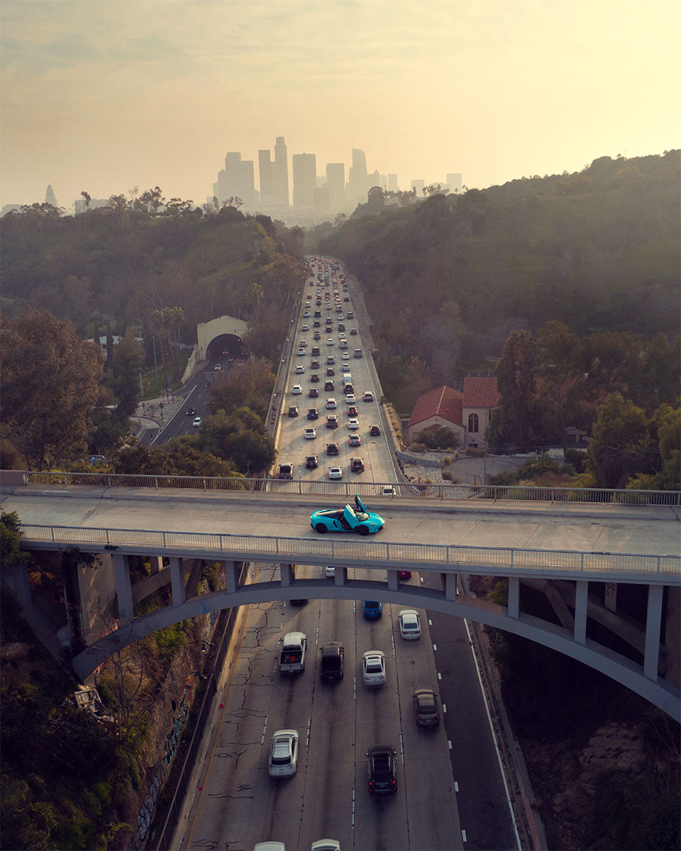 Aerial of a cyan McLaren on an arched bridge above a busy freeway with the Los Angeles skyline in haze, by Niaz Uddin