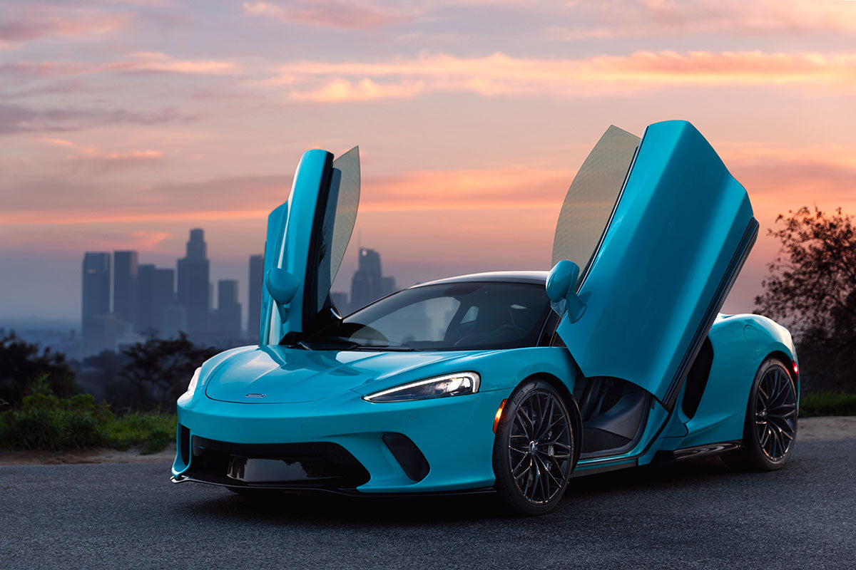 Front three-quarter of a cyan McLaren GT with doors up and the downtown Los Angeles skyline behind at sunset, by Niaz Uddin