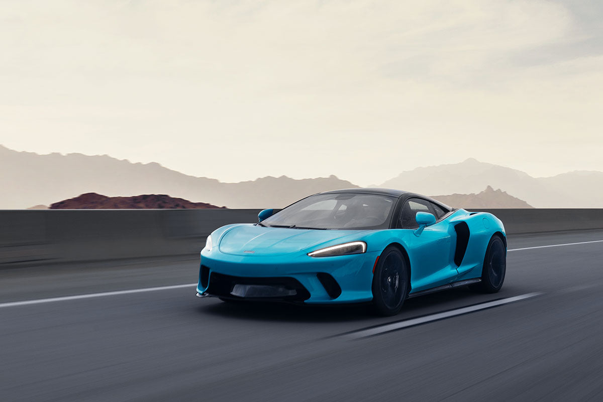 Cyan McLaren GT in motion on an open highway with desert mountains on the horizon, by Niaz Uddin