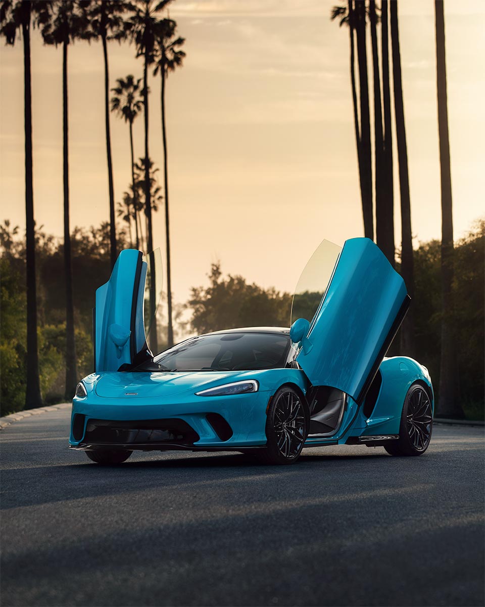 Cyan McLaren GT with doors up on a palm-lined street in golden-hour light, by Niaz Uddin