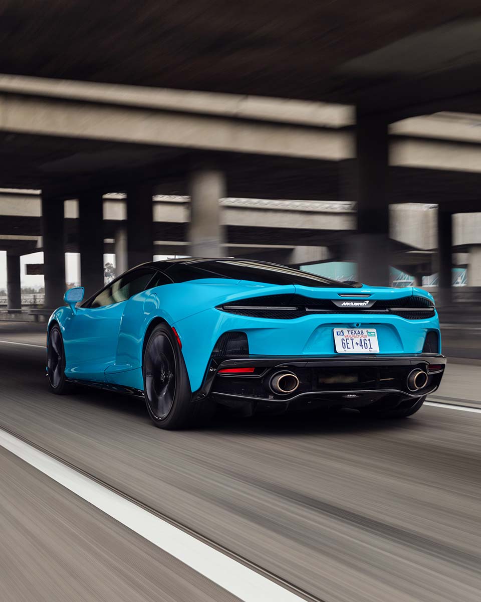 Rear of a cyan McLaren GT in motion under a concrete overpass, by Niaz Uddin
