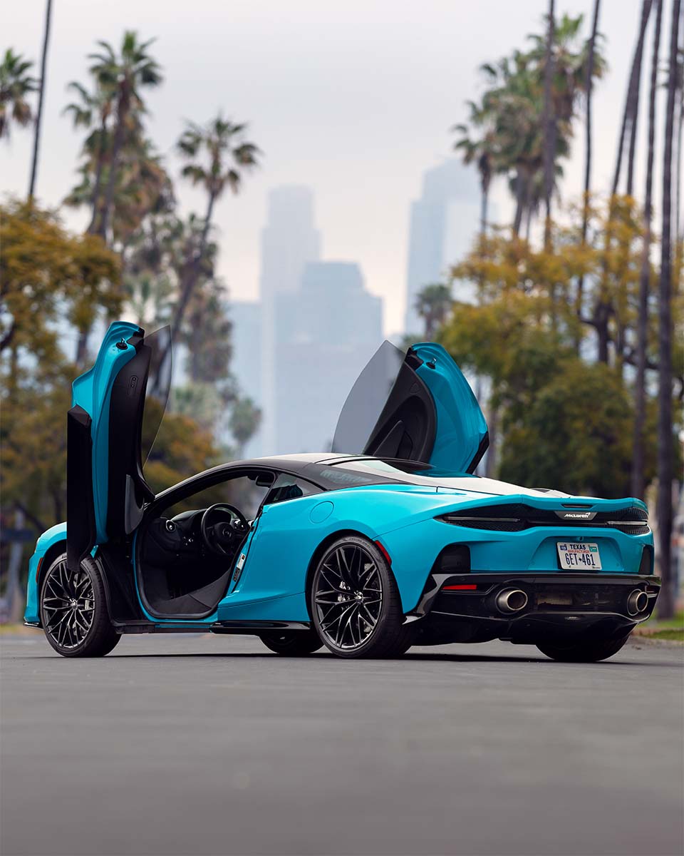 Rear three-quarter of a cyan McLaren GT with doors up on a palm-lined street with the skyline behind, by Niaz Uddin