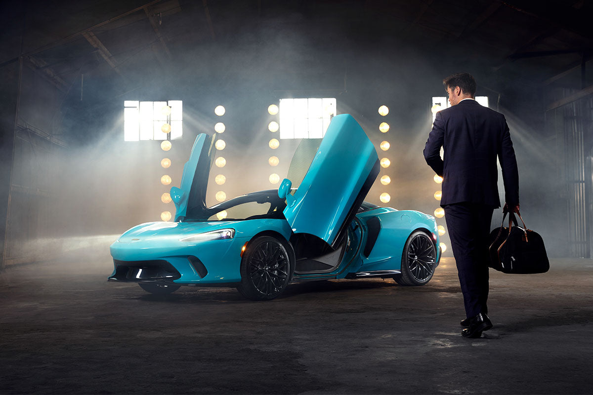 Man in a suit carrying a duffel bag walking toward a cyan McLaren GT with doors up in a moody warehouse, by Niaz Uddin