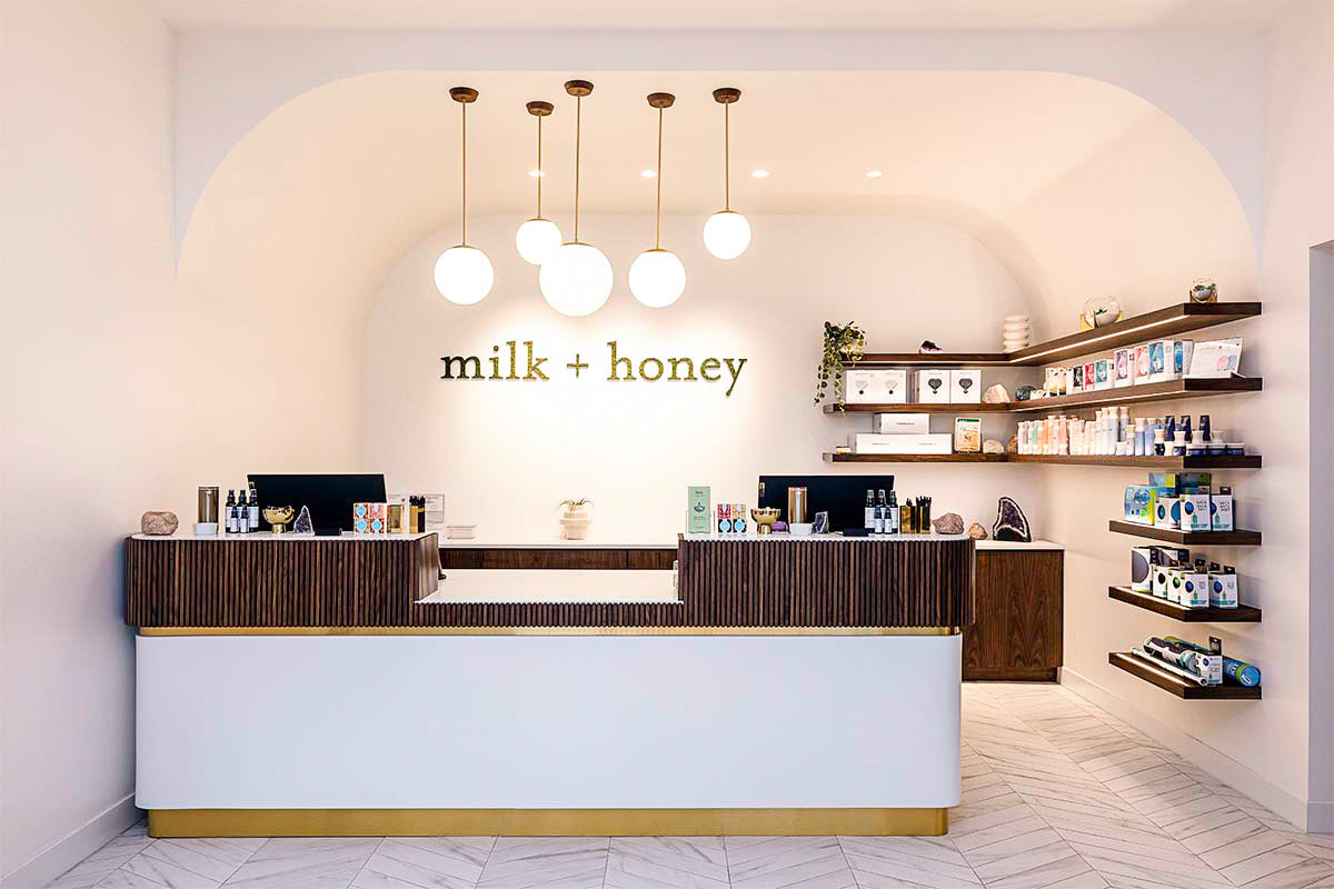 milk + honey spa reception desk with brass globe pendants beneath the branded wall and walnut retail shelving, by Niaz Uddin
