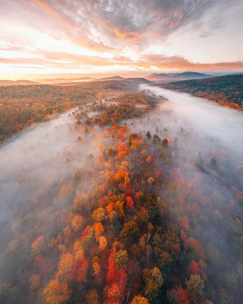 Aerial view at sunrise of peak autumn color along a forest ridge in New York's Adirondack Mountains, low fog rolling through the valley toward distant peaks. Fine art print by Niaz Uddin.