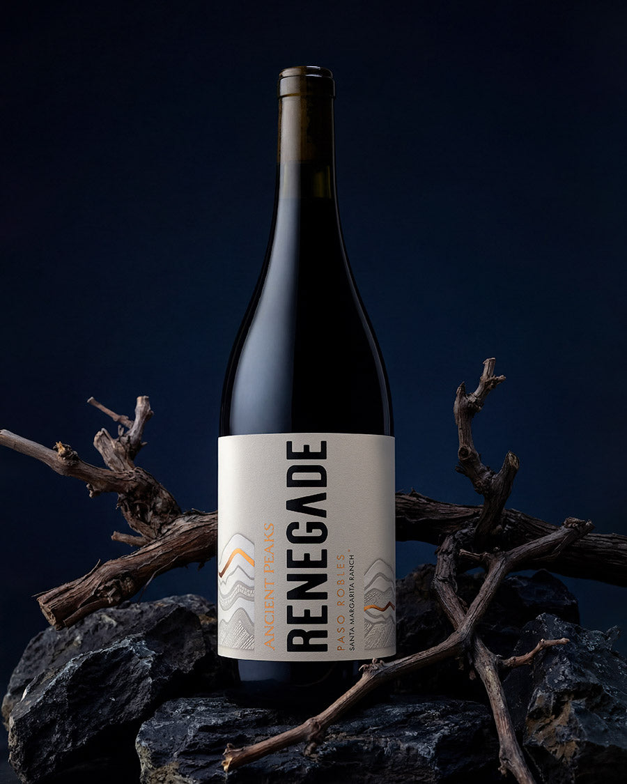 Ancient Peaks Renegade bottle on dark rocks with dried grapevines against a deep blue backdrop, Paso Robles wine photography by Niaz Uddin