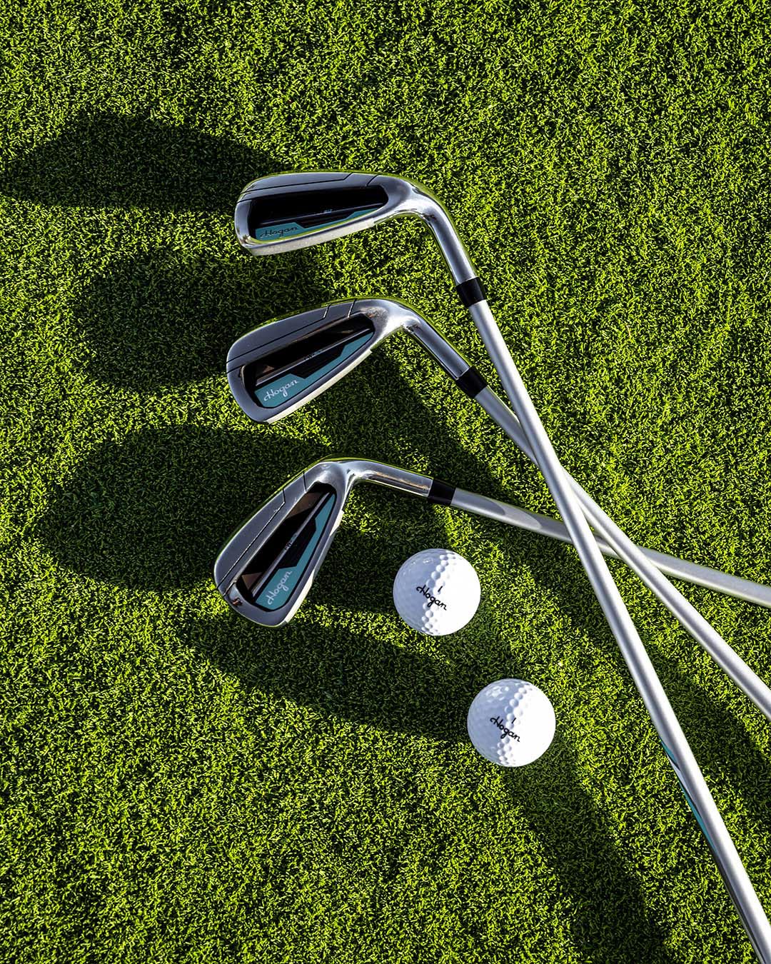 Flat-lay of three Ben Hogan irons and two golf balls on turf with hard shadows, by Niaz Uddin