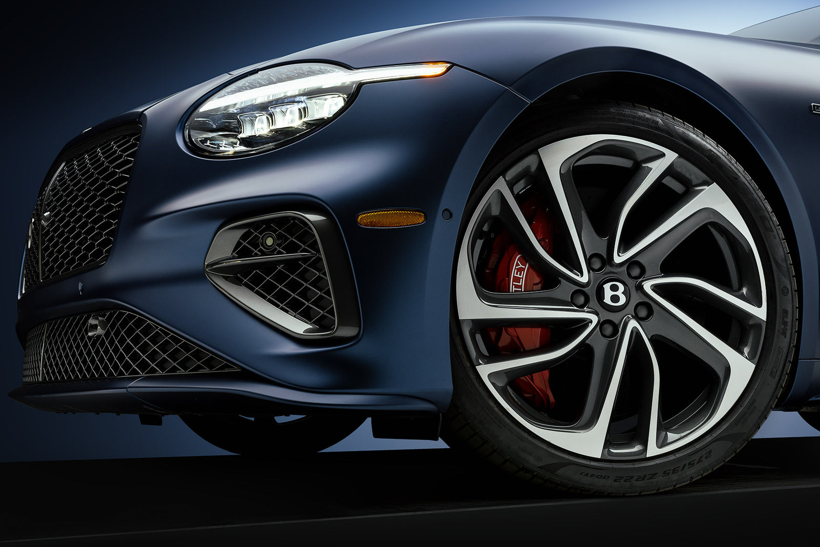 Close detail of a satin blue Bentley Continental GT front wheel, red caliper, and headlight in studio light, by Niaz Uddin