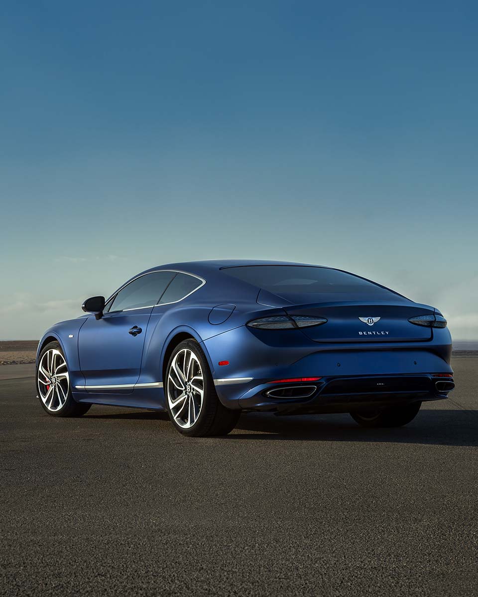 Rear three-quarter of a satin blue Bentley Continental GT on open tarmac by the coast, by Niaz Uddin