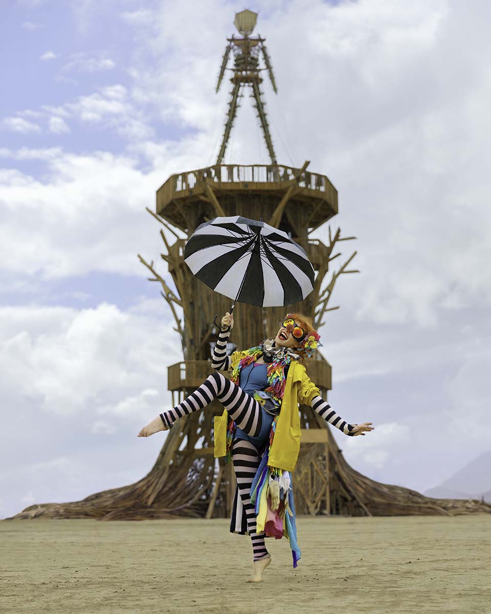 Bonnie Morgan in a striped clown costume with a parasol leaping in front of the Man, Burning Man 2026, by Niaz Uddin
