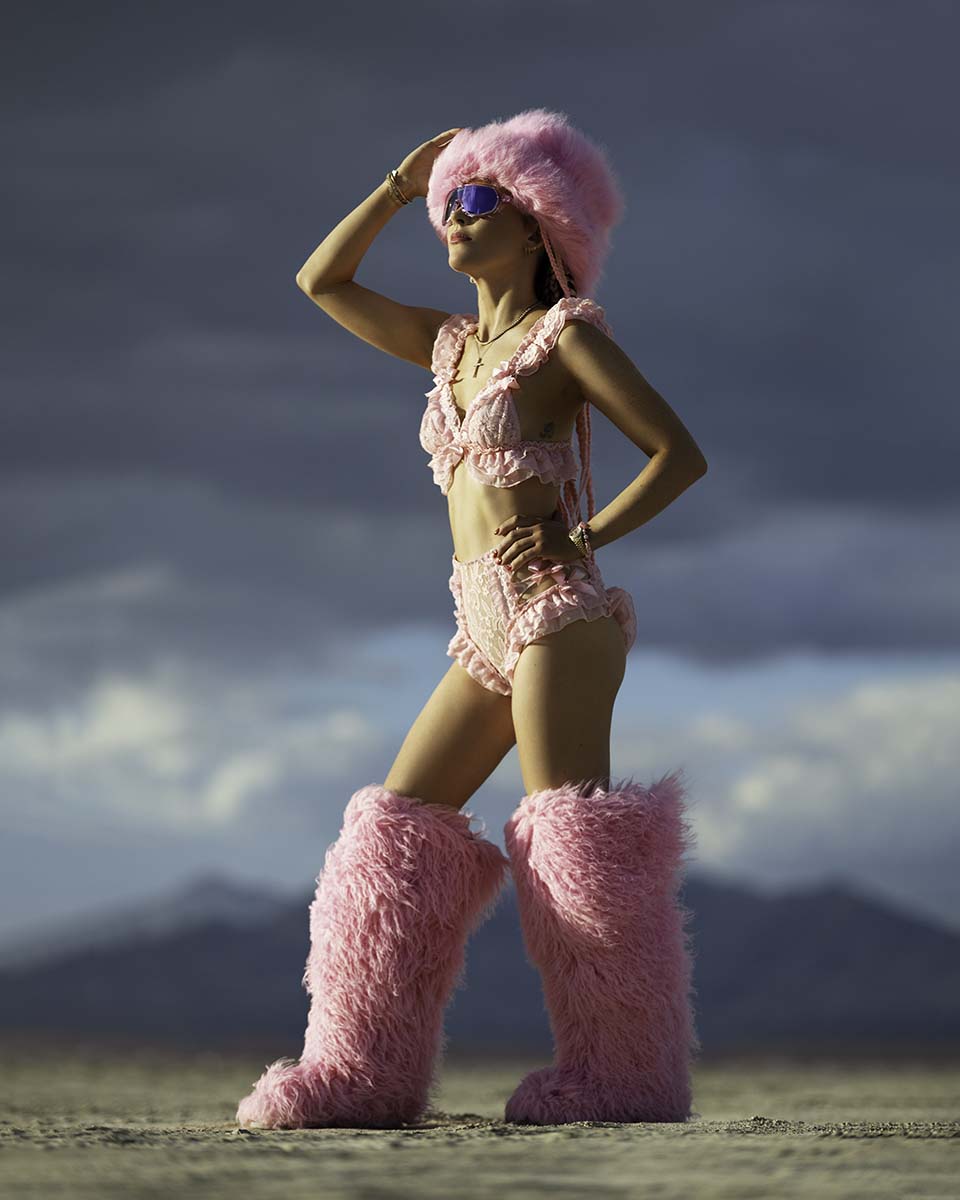 Nicole Renzulli in a pink fur hat and boots posing on the playa under storm clouds, Burning Man 2026, by Niaz Uddin