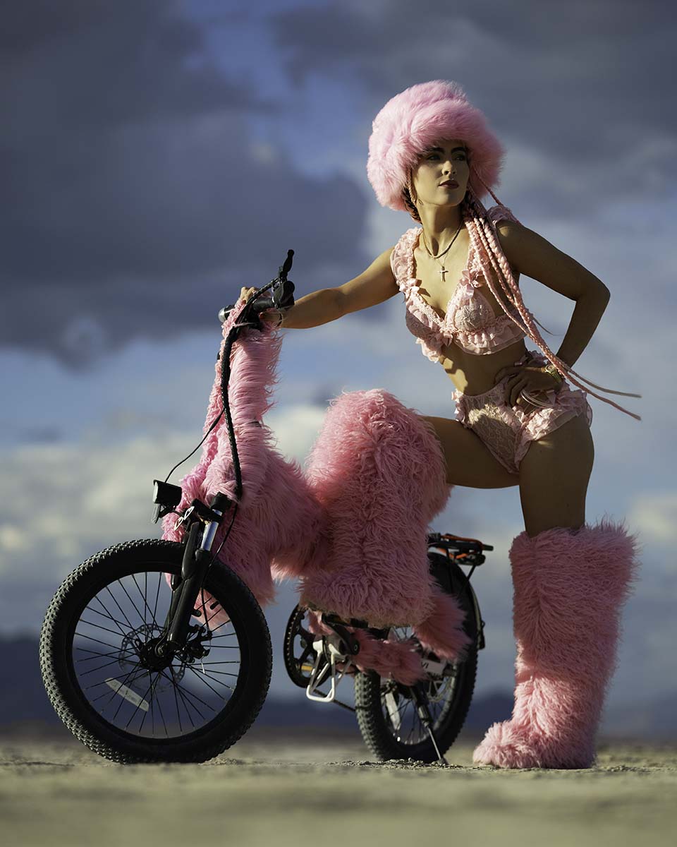 Nicole Renzulli in pink fur posing with a matching pink fur bike under a stormy sky, Burning Man 2026, by Niaz Uddin