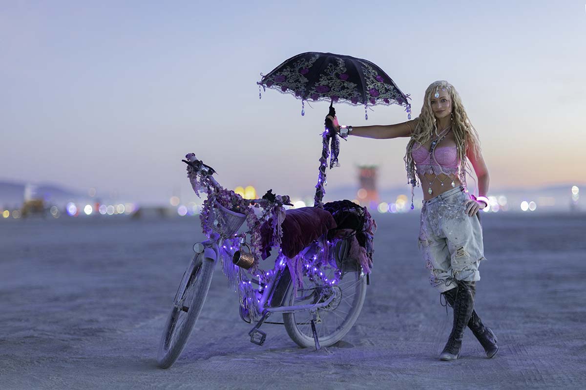 Mindy Robinson holding a lace parasol beside a decorated bicycle on the playa at dusk, Burning Man 2026, by Niaz Uddin