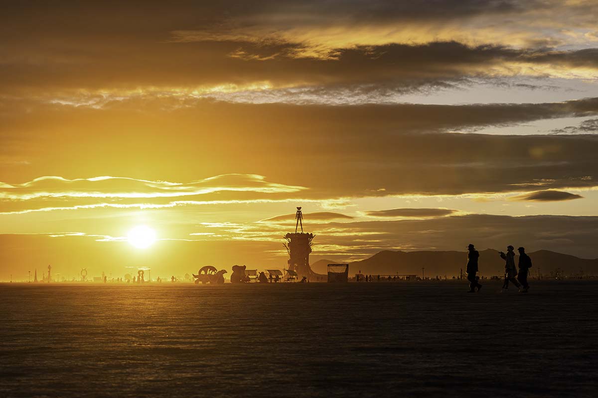 Silhouette of the Man against a golden sunrise over the playa with distant figures and art cars, Burning Man 2026, by Niaz Uddin