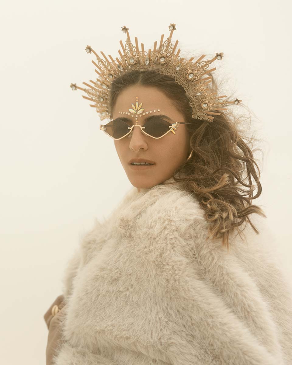 Vanessa Escote in a gold filigree crown, face jewels and a cream fur coat, glancing over her shoulder in a whiteout dust storm, Burning Man 2026, by Niaz Uddin