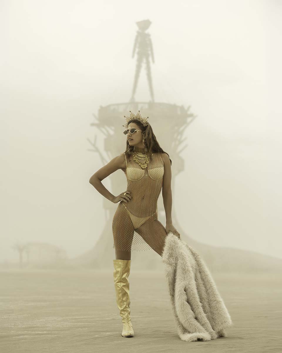Vanessa Escote in gold with a fur wrap in a dust storm, the Man faint behind her, Burning Man 2026, by Niaz Uddin