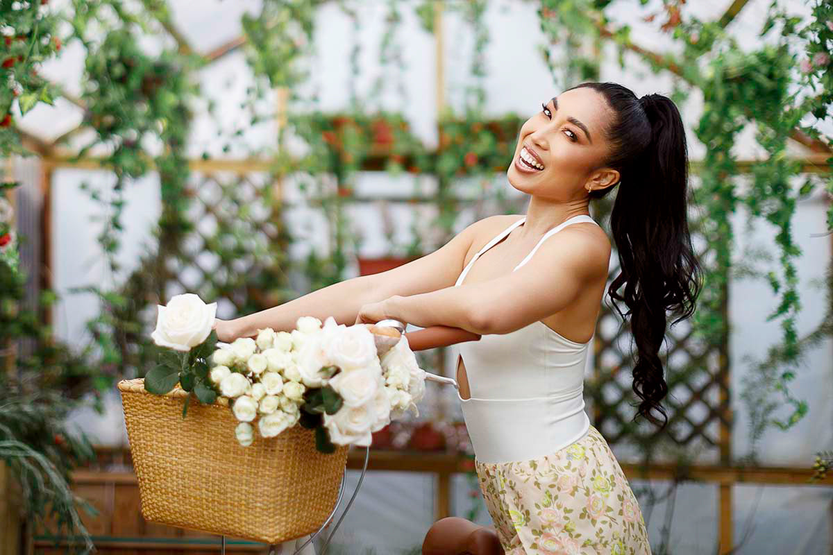 Cassey Ho laughing on a bicycle with a basket of white roses in a rose-covered greenhouse, by Niaz Uddin