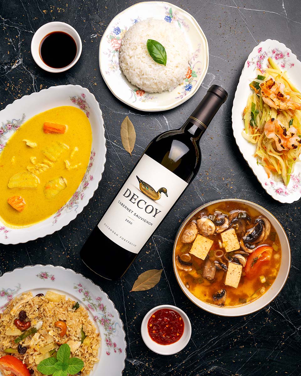 Decoy Cabernet Sauvignon at the center of a Thai feast with curry, rice, soup, fried rice, and papaya salad on floral china, by Niaz Uddin