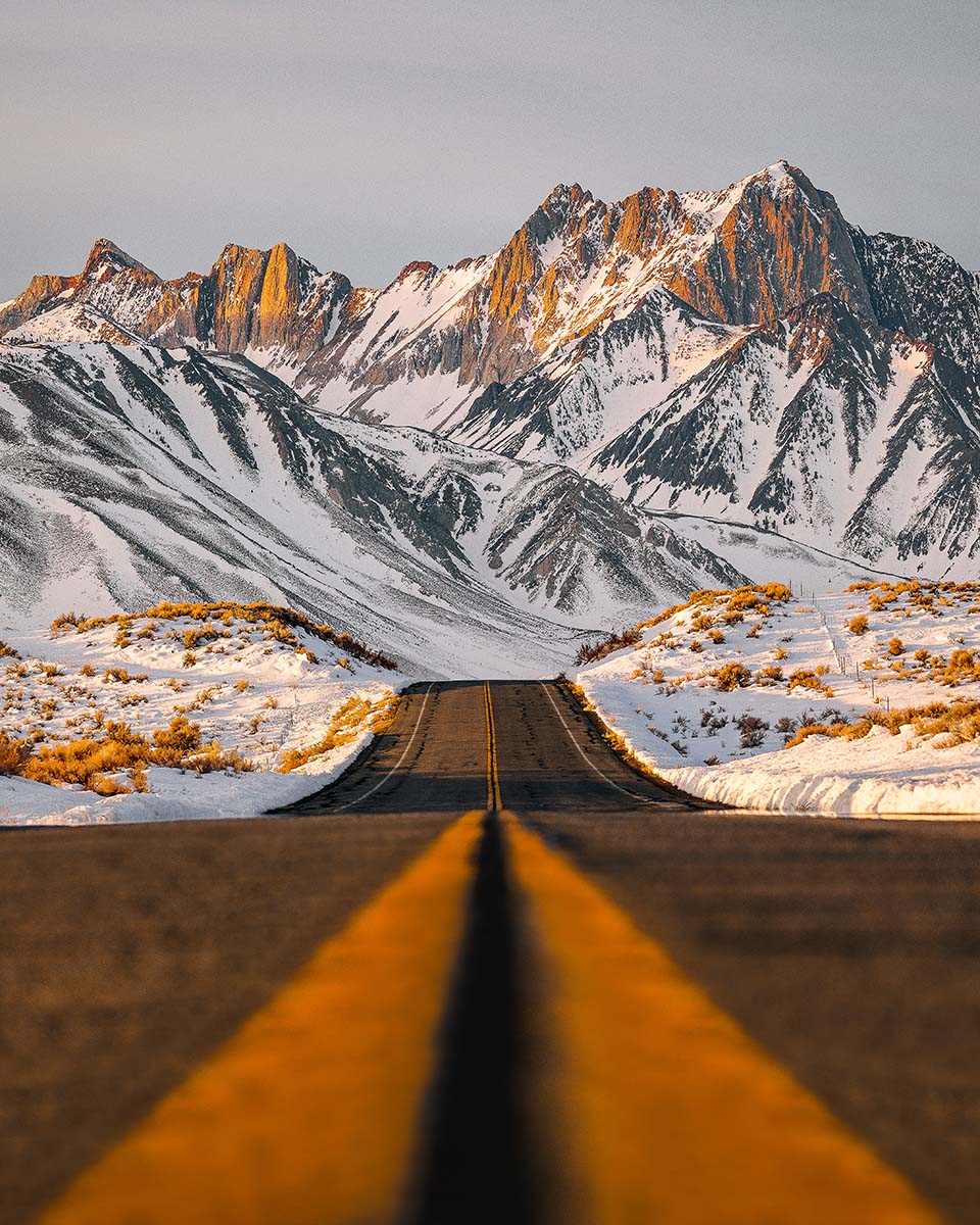 A two-lane road leading toward snow-covered Eastern Sierra peaks lit by sunrise alpenglow at Benton Crossing near Mammoth, a California fine art print by Niaz Uddin.