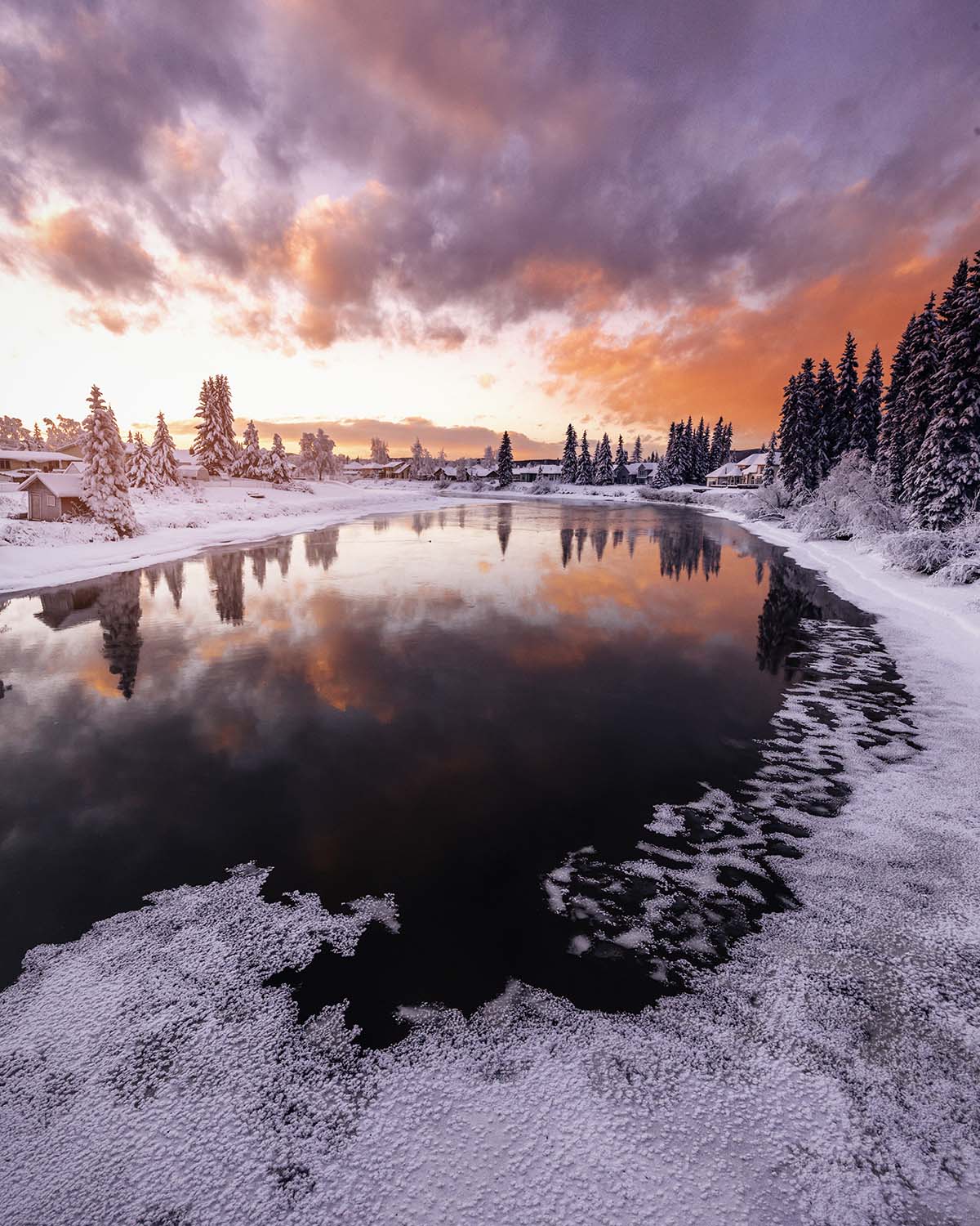 Winter sunset on the half-frozen Chena River near Fairbanks, Alaska, a fiery pink and orange sky above snowy spruce with frost feathers spreading across dark reflecting water. Fine art print by Niaz Uddin.