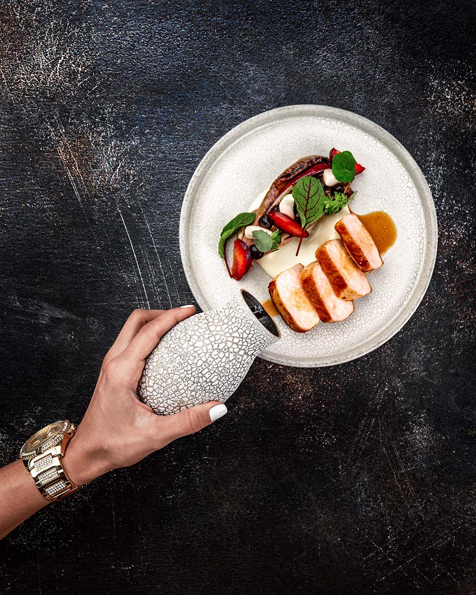Sliced roast pork loin with plums and greens on a speckled plate as a hand pours sauce from a textured jug, by Niaz Uddin