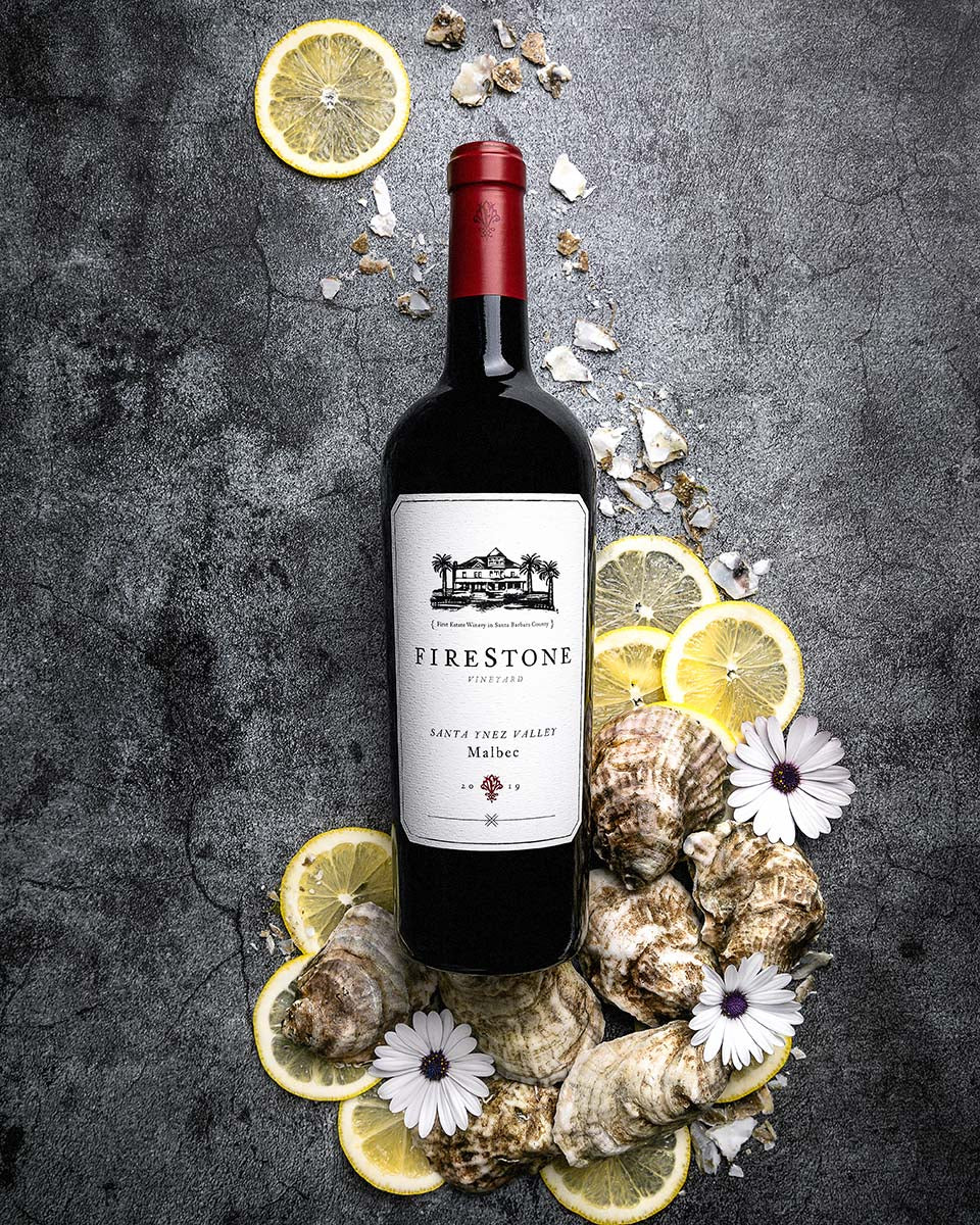 Firestone Vineyard Malbec surrounded by oyster shells, lemon slices, and white daisies on dark slate, by Niaz Uddin