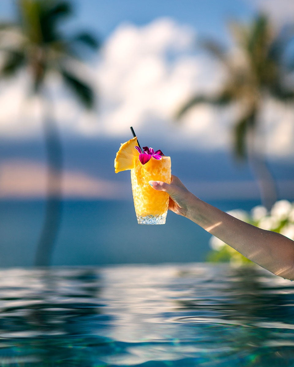 Hand holding a pineapple-and-orchid tropical cocktail against blurred palms and the ocean at dusk, Four Seasons Resort Maui, by Niaz Uddin