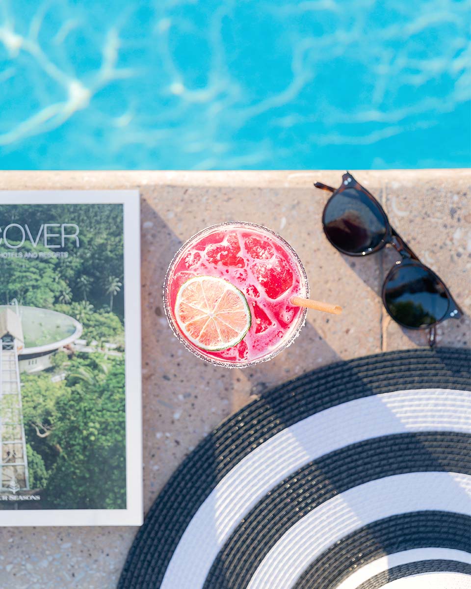 Overhead poolside flatlay of a pink cocktail, sunglasses, sun hat, and a Four Seasons Discover magazine, by Niaz Uddin