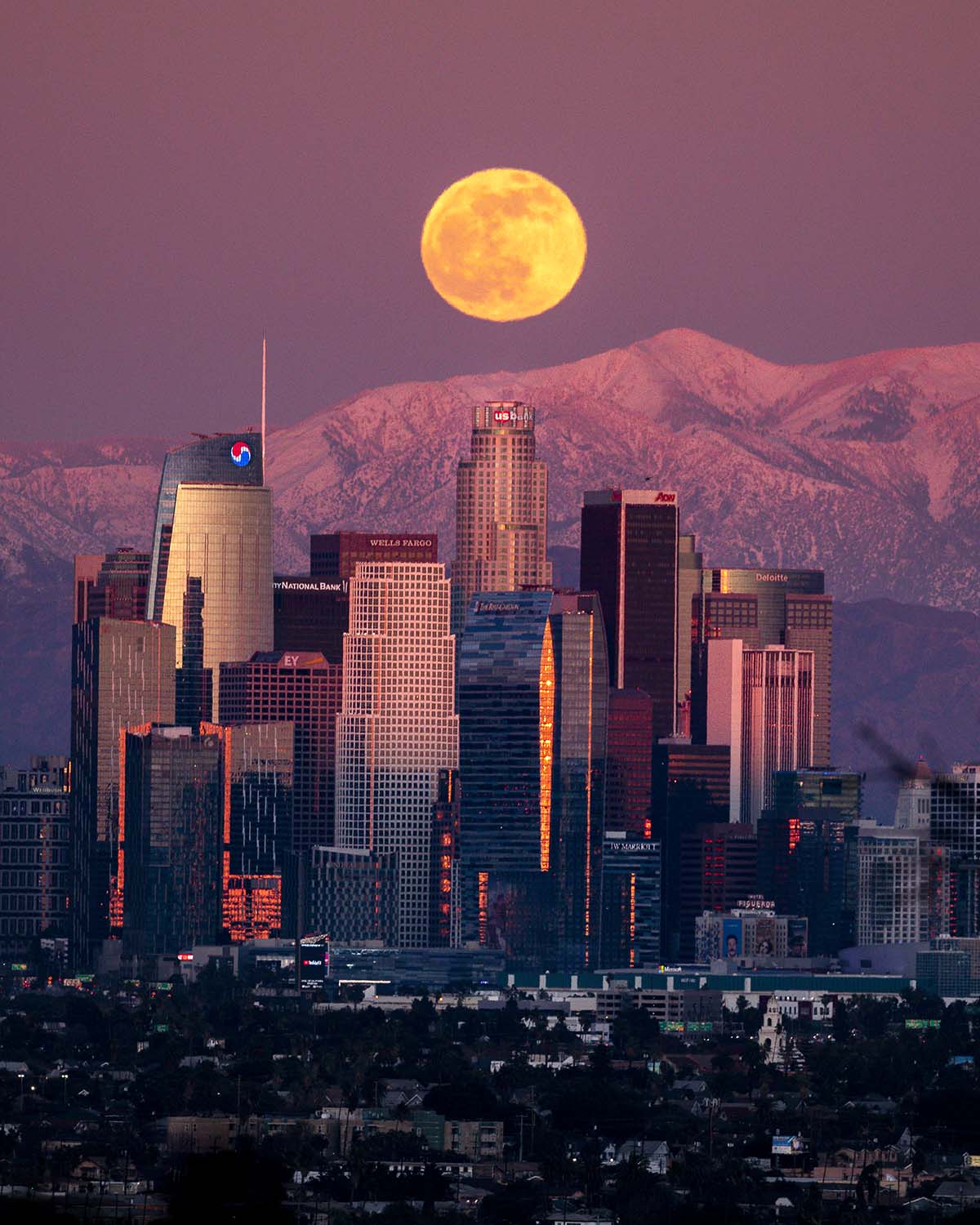 A full moon rising over the downtown Los Angeles skyline with the snow-capped San Gabriel Mountains at twilight.
