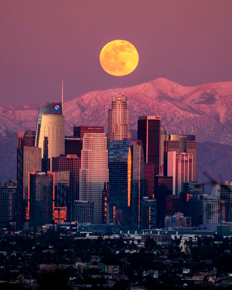 A full moon rising over the downtown Los Angeles skyline with the snow-capped San Gabriel Mountains at twilight.