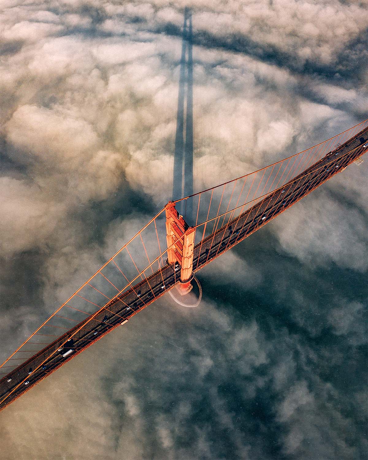 An aerial view looking down on the north tower of the Golden Gate Bridge breaking through low fog at sunrise, its long shadow cast across the clouds, a San Francisco fine art print by Niaz Uddin.