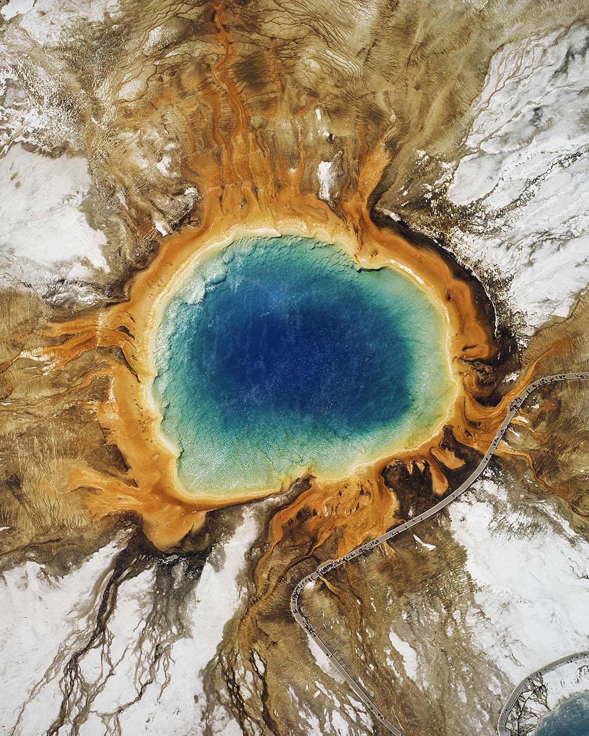 An aerial view looking straight down on Grand Prismatic Spring in Yellowstone, its deep blue center ringed by orange bacterial mats and surrounding snow, a Wyoming fine art print by Niaz Uddin.