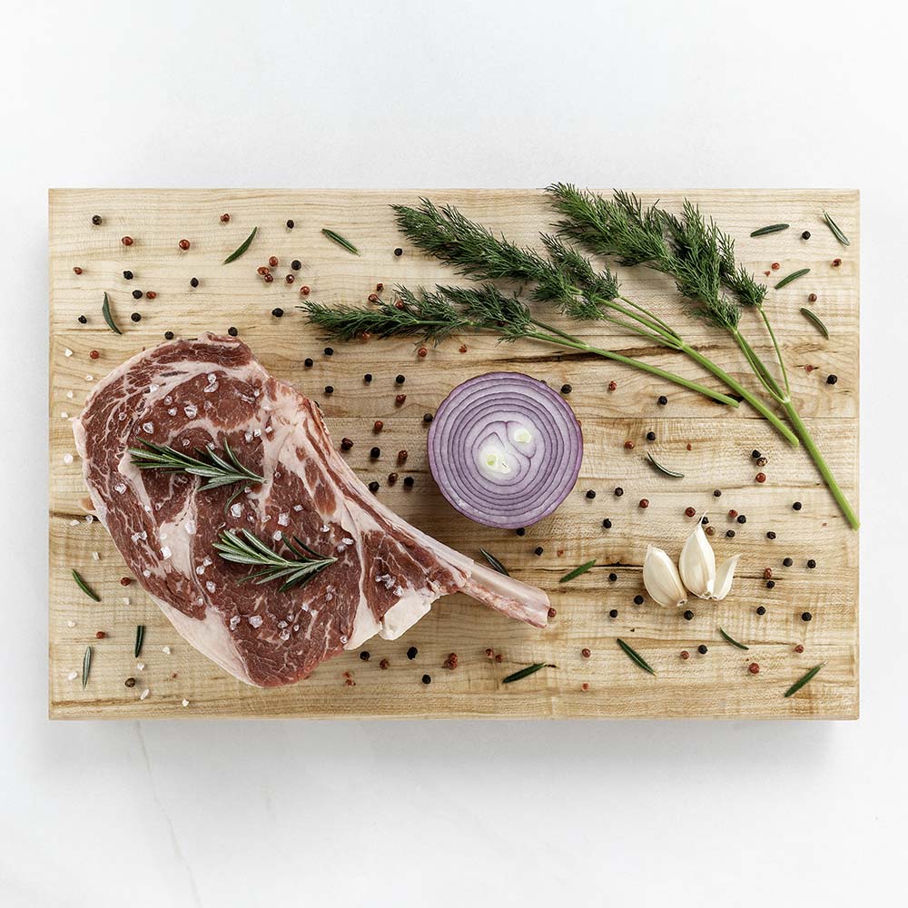 Tomahawk ribeye steak with rosemary, sea salt, red onion, and peppercorns on a Hawker Woodcraft maple cutting board, by Niaz Uddin