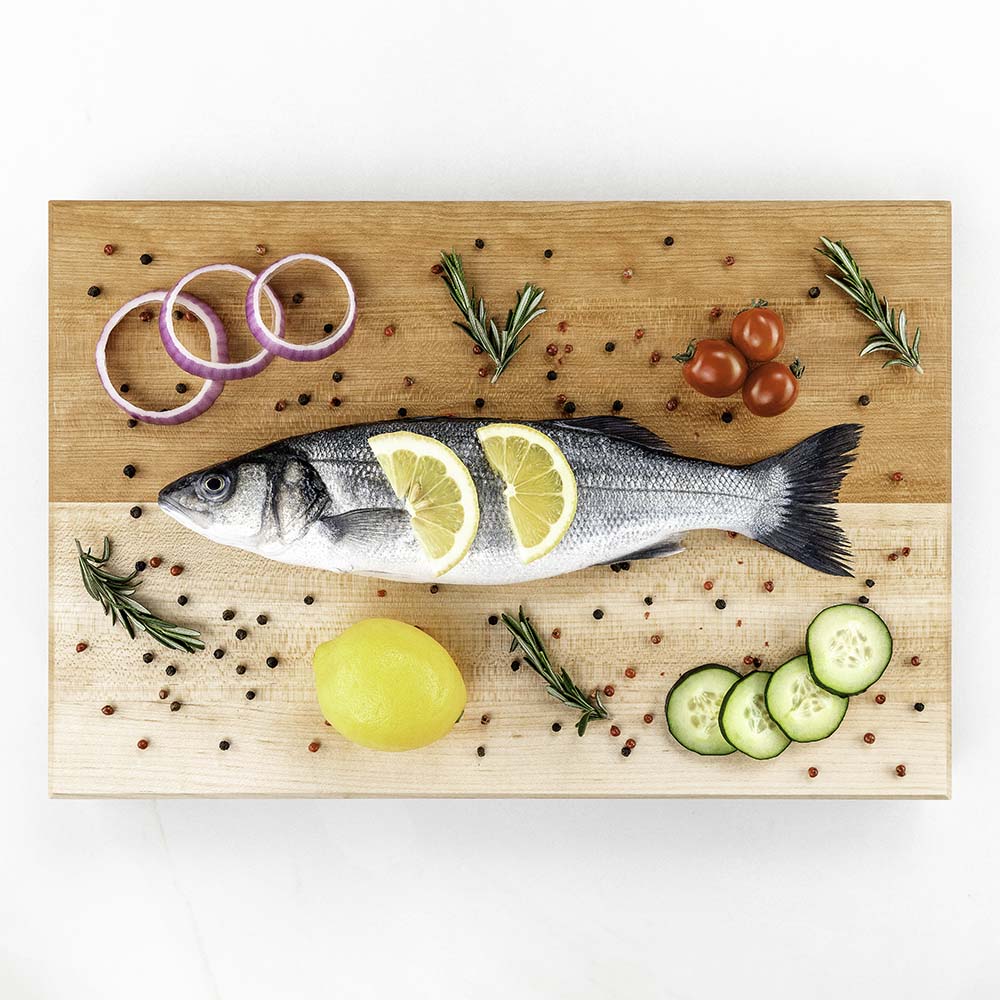 Whole sea bass with lemon slices, red onion, cherry tomatoes, and cucumber styled on a Hawker Woodcraft maple serving board, by Niaz Uddin