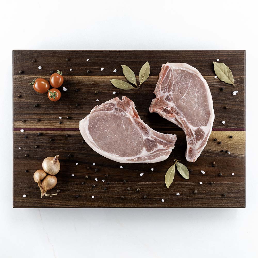 Two bone-in pork chops with bay leaves, peppercorns, sea salt, and shallots on a Hawker Woodcraft walnut cutting board, by Niaz Uddin