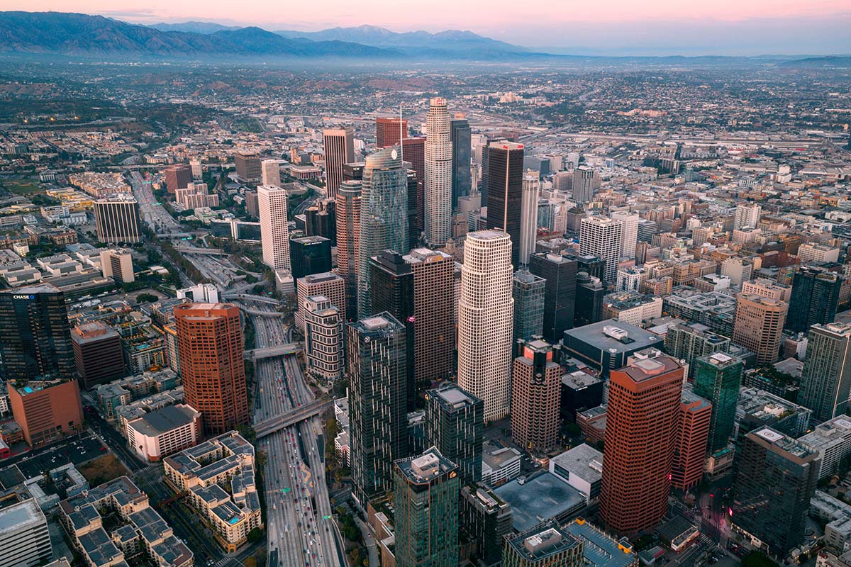 Wide aerial of downtown Los Angeles at golden hour with the USC tower in the foreground and the San Gabriel Mountains behind, by Niaz Uddin