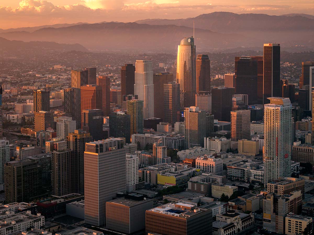 Wide aerial of the downtown Los Angeles skyline backed by the San Gabriel Mountains under a pink sky at golden hour, by Niaz Uddin