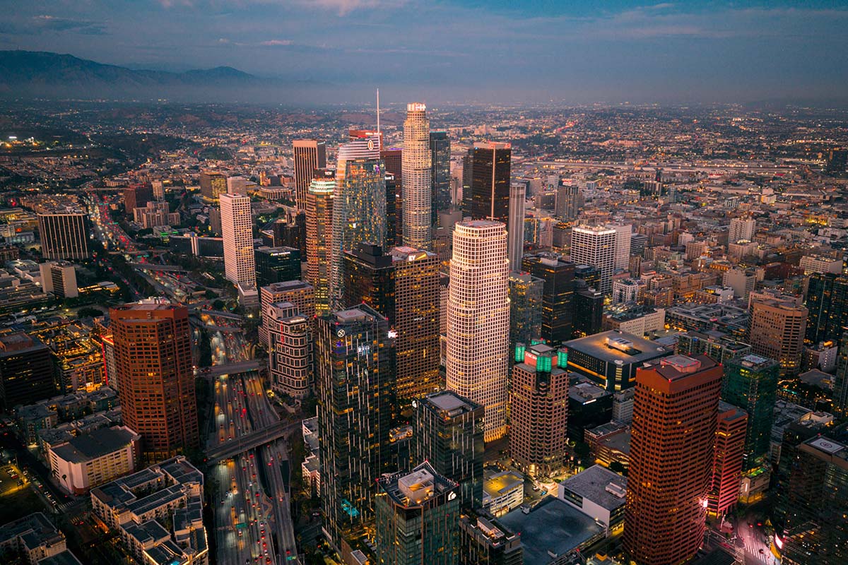 Symmetrical aerial straight down the downtown Los Angeles corridor toward the towers at sunset under a pink sky, by Niaz Uddin