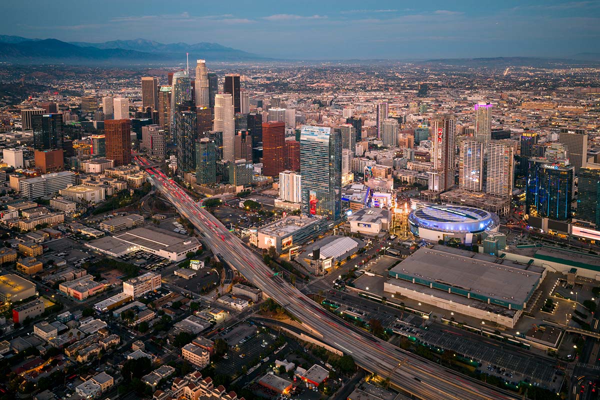 Wide twilight aerial of the downtown Los Angeles skyline and surrounding city under a soft pink sky, by Niaz Uddin