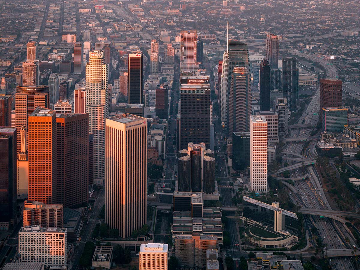 Telephoto aerial of downtown Los Angeles towers compressed against the San Gabriel Mountains at golden hour, by Niaz Uddin