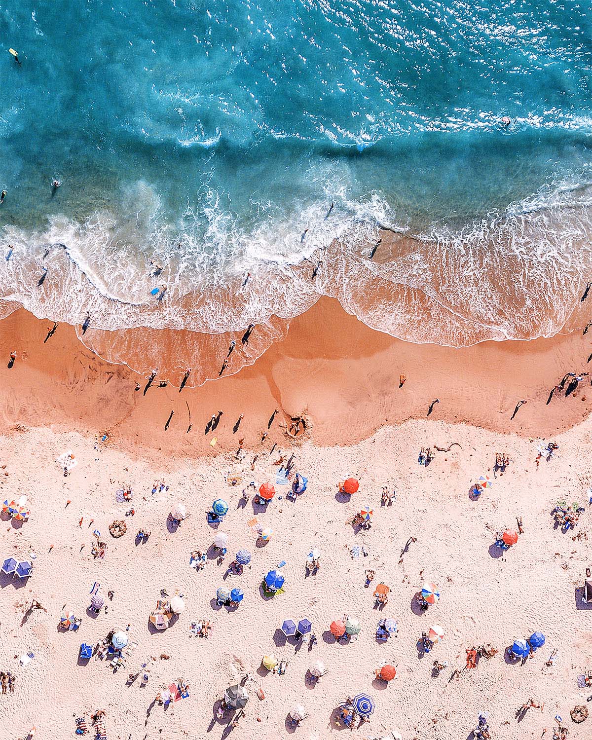 Aerial view straight down on Laguna Beach, California at peak summer, colorful umbrellas scattered across pink sand with a wave breaking in a long white curl. Fine art print by Niaz Uddin.