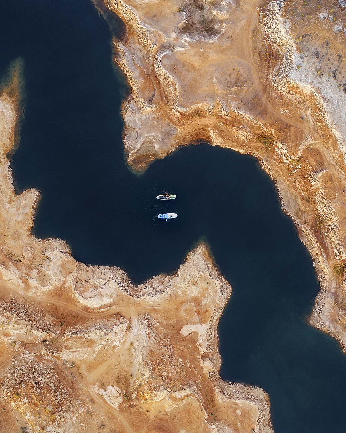 Aerial view straight down on two paddleboarders on the deep blue water of Lake Powell, surrounded by pale sandstone shoreline exposed by falling water levels. Fine art print by Niaz Uddin.