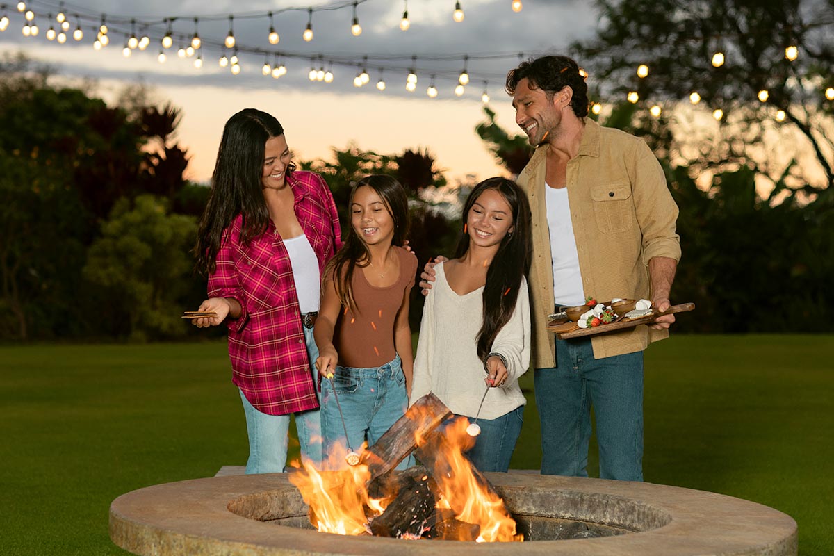 Family roasting s'mores around a fire pit under string lights at sunset at Makena Beach Club, Maui, by Niaz Uddin