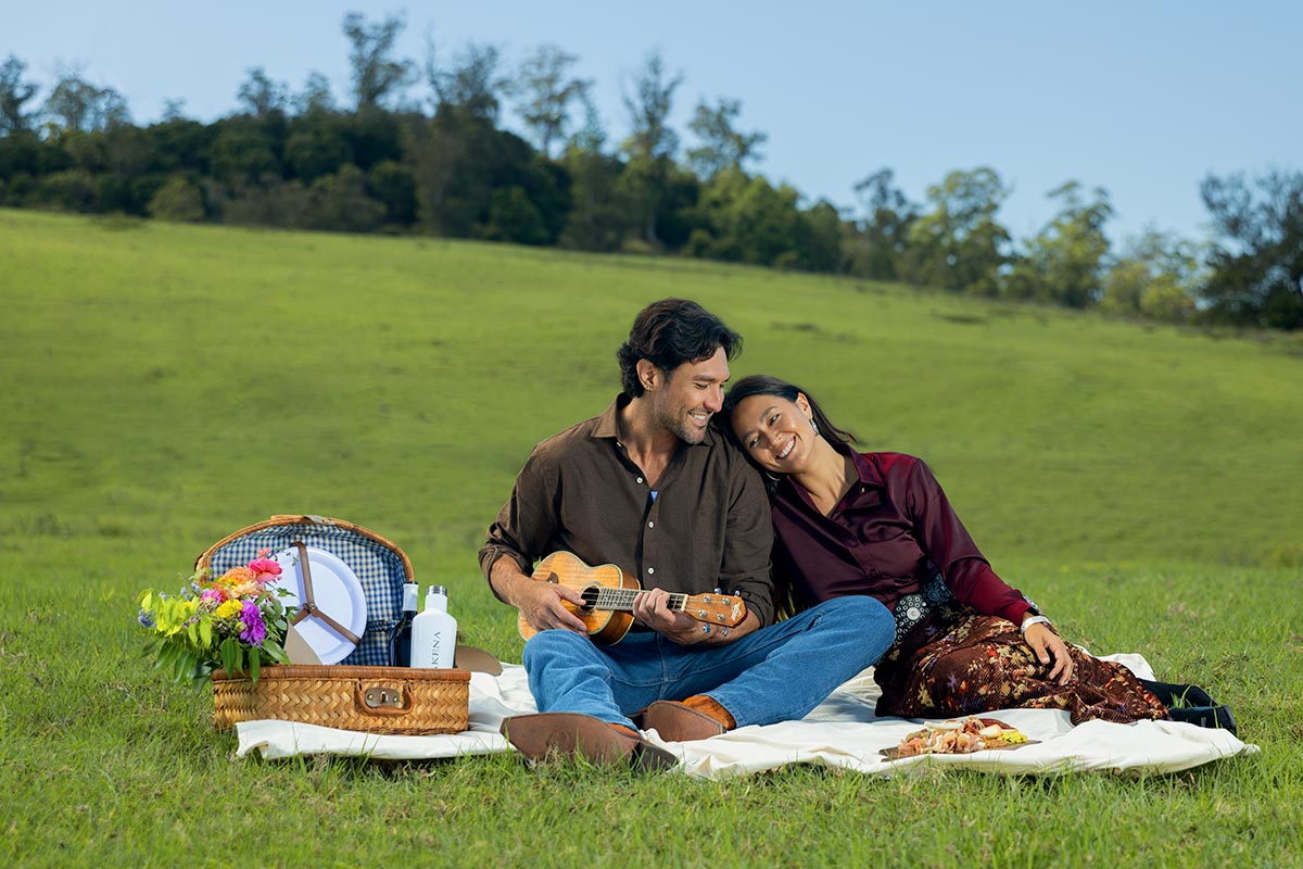 Couple on a picnic blanket in a green meadow, one playing ukulele, at Makena Beach Club, Maui, by Niaz Uddin