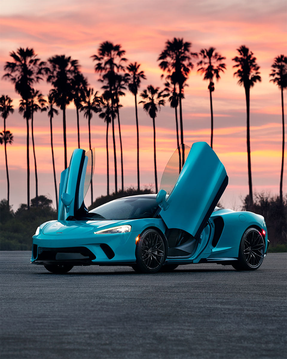 Cyan blue McLaren GT with dihedral doors open against a row of palm trees at pink sunset, by Niaz Uddin