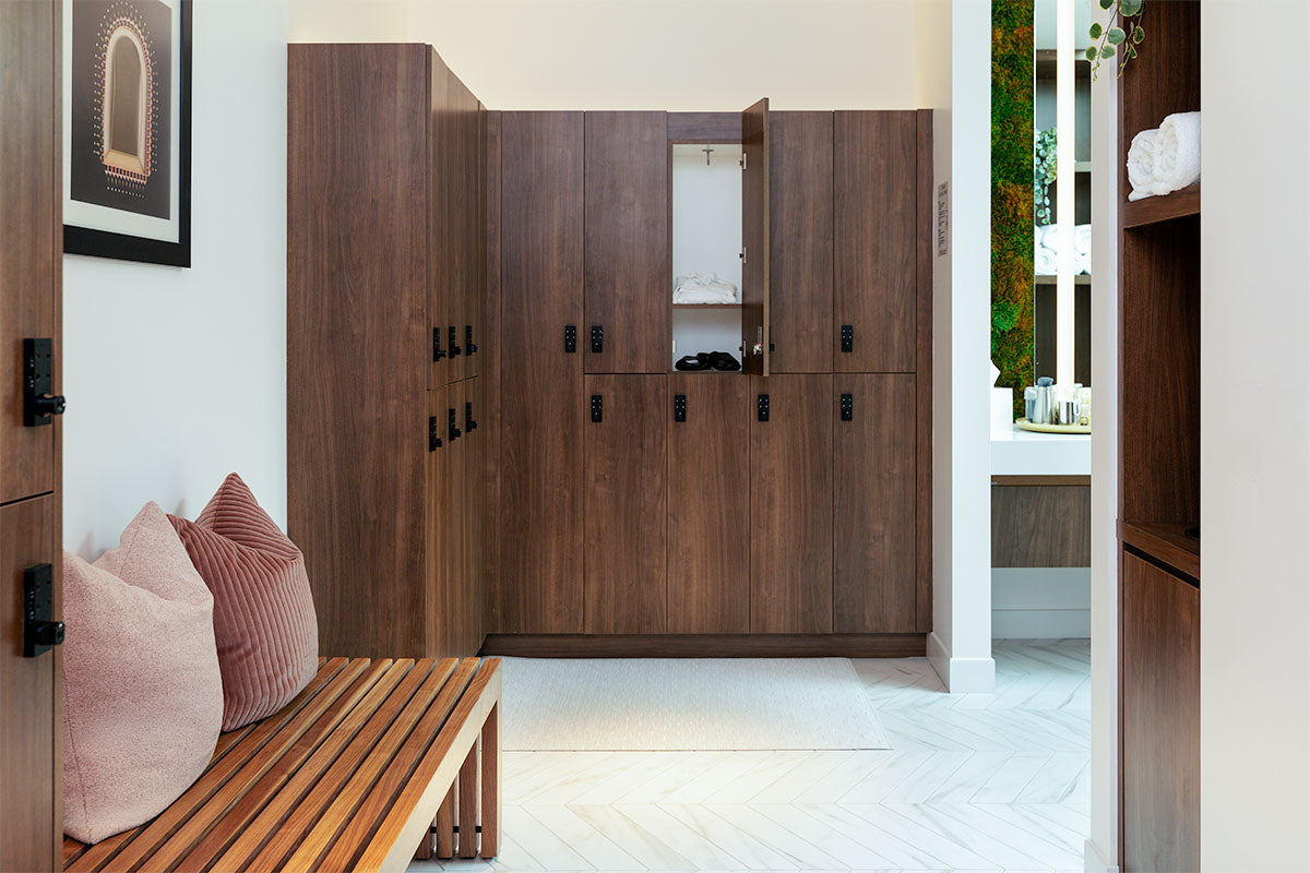 milk + honey locker room with a slatted teak bench, dusty-rose pillows, and walnut lockers, one open with robe and slippers, by Niaz Uddin