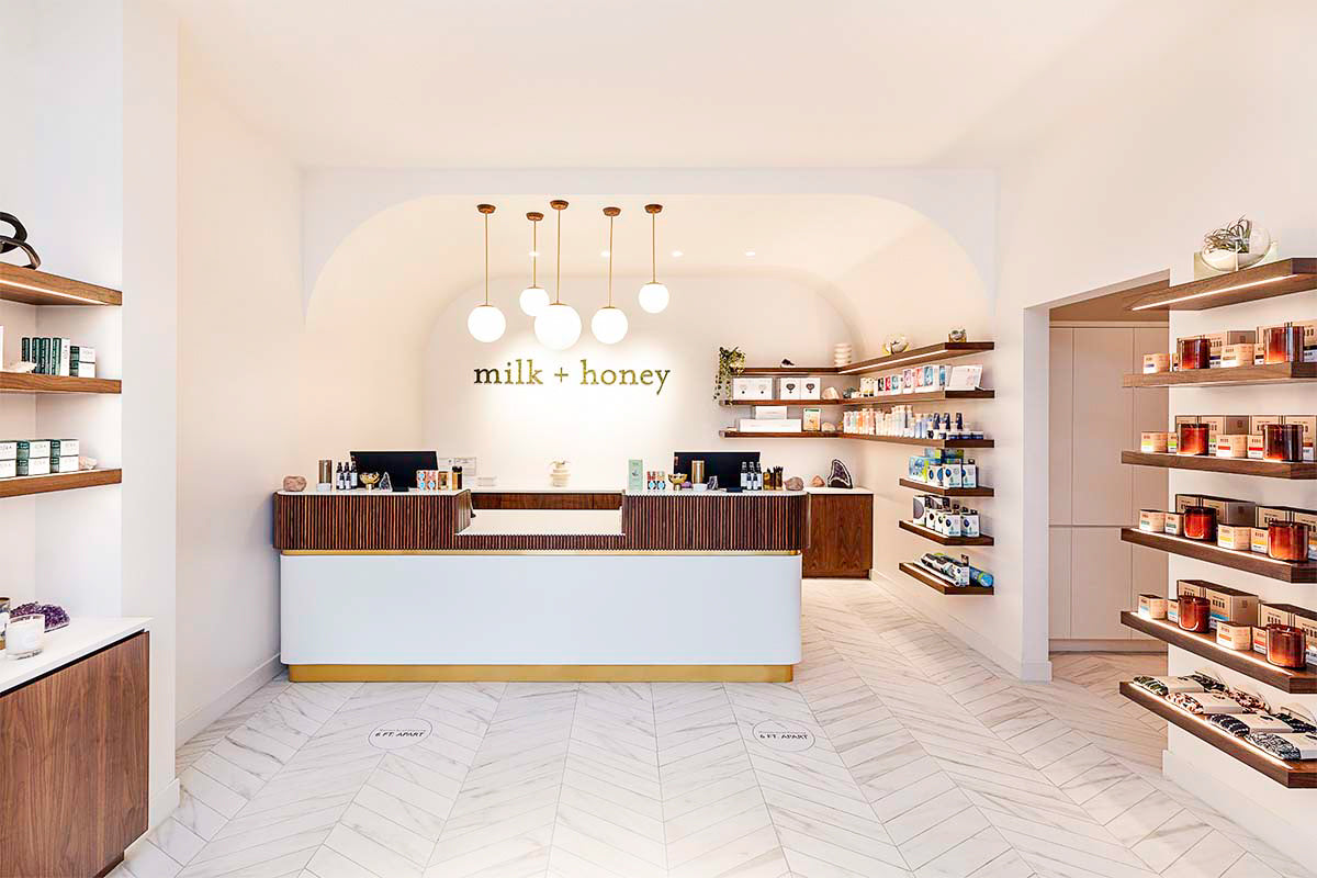 milk + honey reception and retail floor seen wide, with brass globe pendants, the branded wall, and walnut shelving, by Niaz Uddin