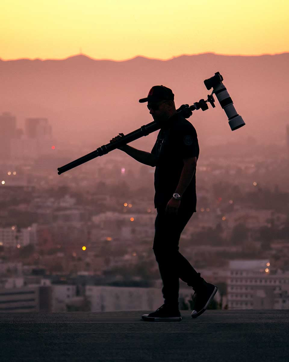 Niaz Uddin carrying a tripod and telephoto lens above the Los Angeles skyline at sunset