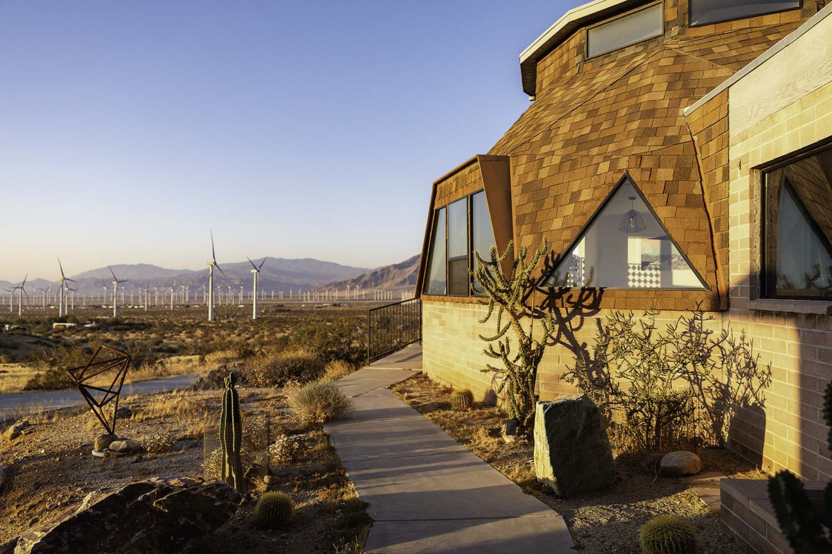 Side view of the shingled geodesic dome house with a triangular window and walkway, wind turbines and desert mountains beyond at golden hour, by Niaz Uddin